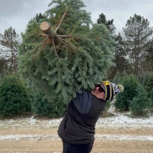 Customer holding a freshly cut Christmas tree above her head with tree fields in the background.